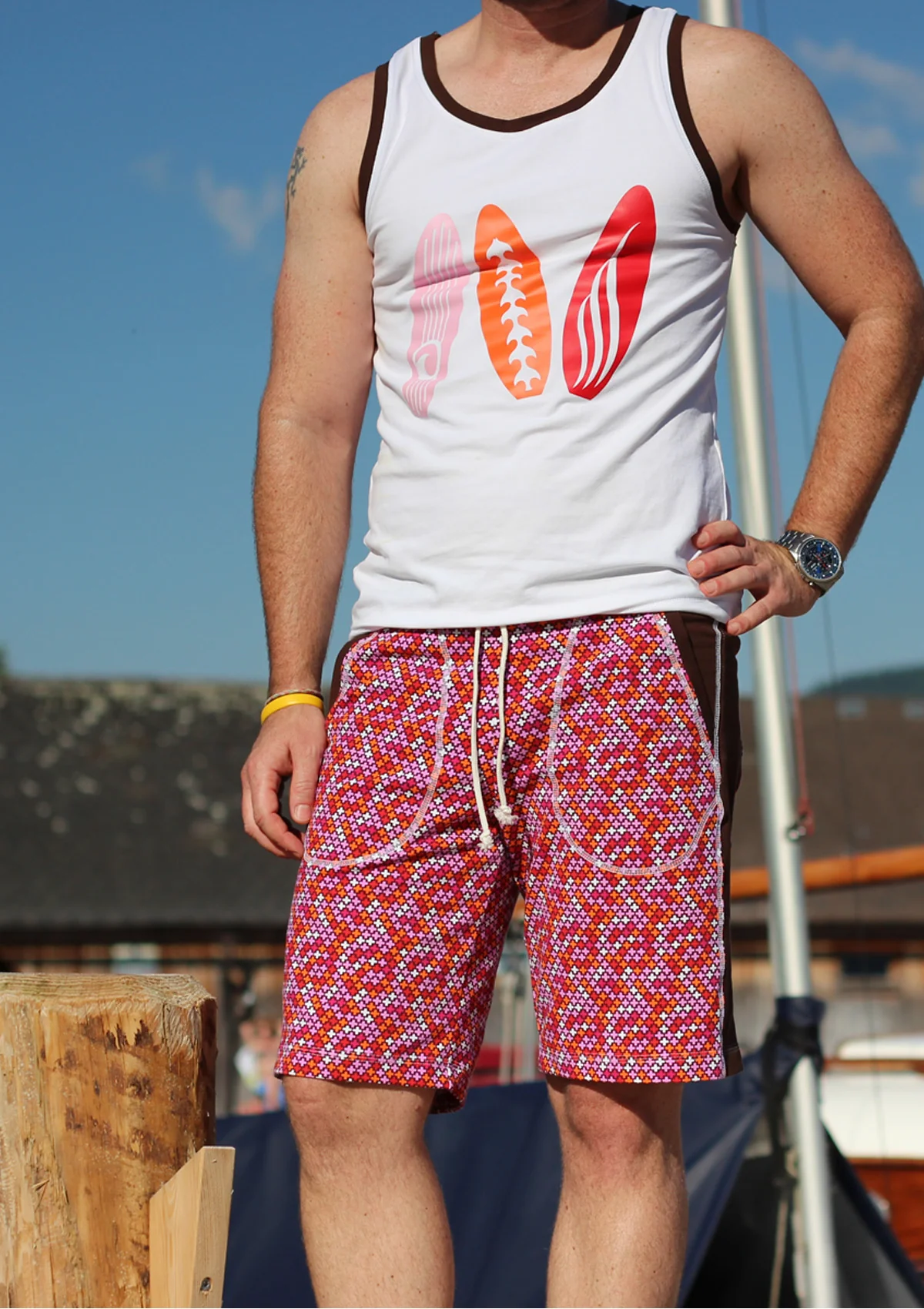 Man wearing a white sleeveless tank top with brown trim and three colorful surfboard graphics, paired with bright pink patterned swim shorts, standing outdoors on a sunny day.