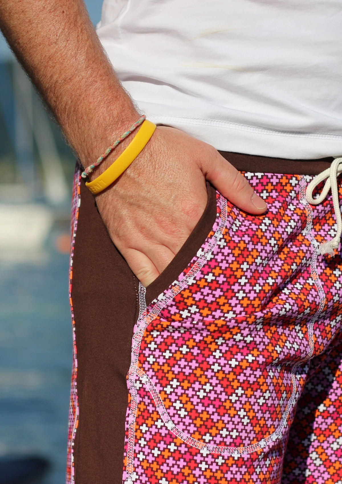 Close-up of a man's hand in a pocket of colorful patterned swim shorts, with a yellow bracelet on the wrist.