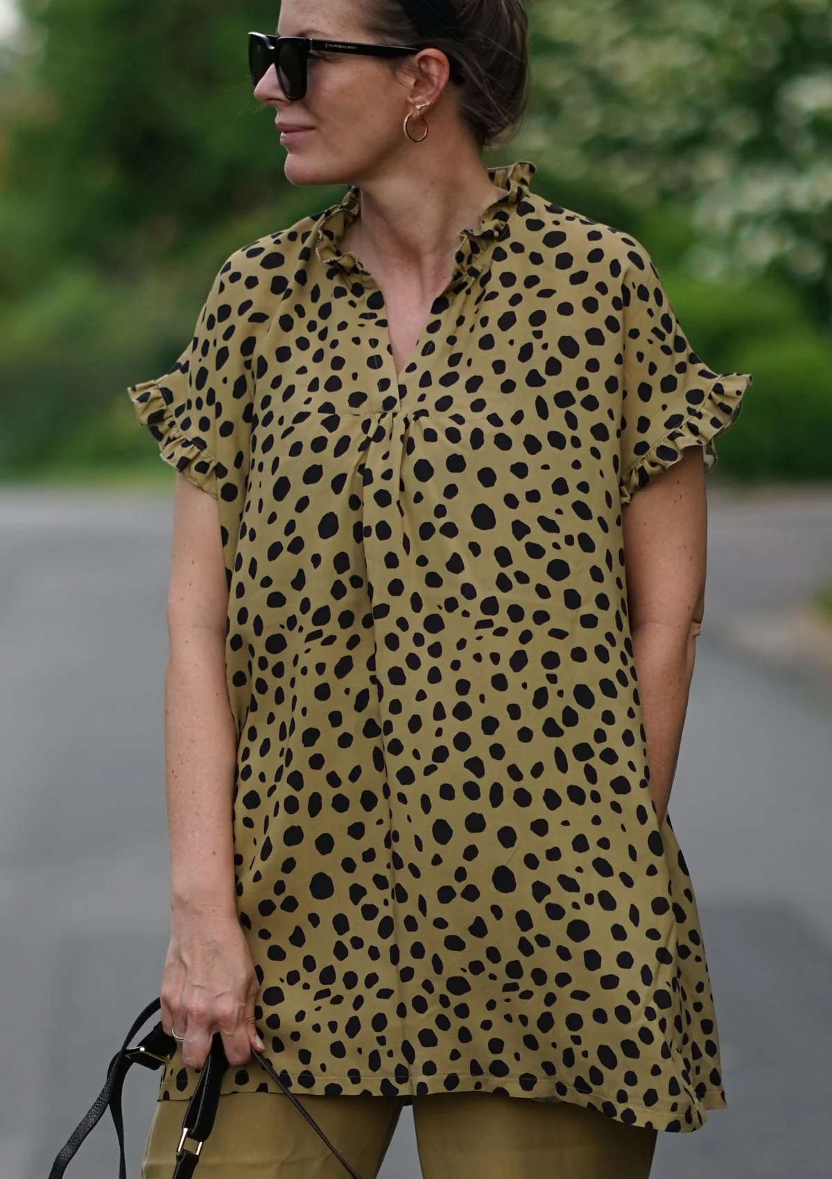 Side profile of a woman wearing a tan leopard-print blouse with ruffled sleeves, black sunglasses, and gold hoop earrings outdoors.